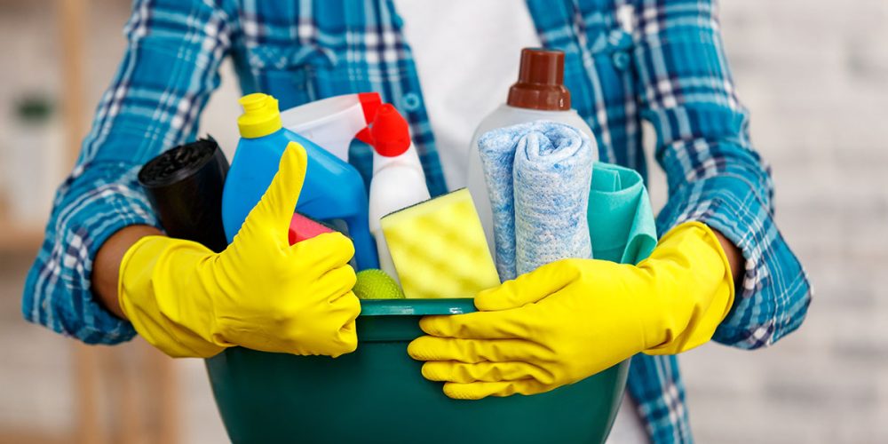 Studio shot of housekeeper while cleaning room. Woman wearing gloves, showing thumb up and holding bowl full of bottles with disinfectant